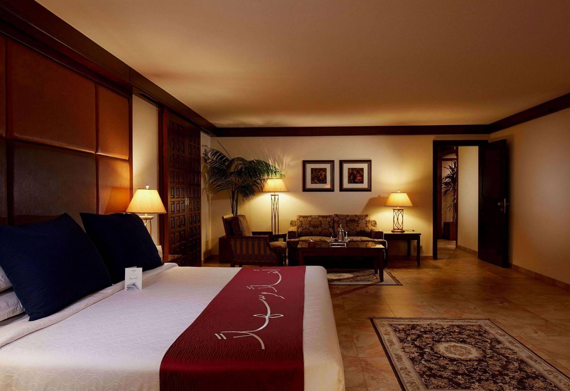 Makarem Ajyad Makkah Hotel Мекка Экстерьер фото The photo depicts a cozy hotel room with a warm and inviting atmosphere. It features a large bed with dark blue pillows and a maroon throw blanket. On either side of the bed, there are lamps providing soft lighting. In the background, a small seating