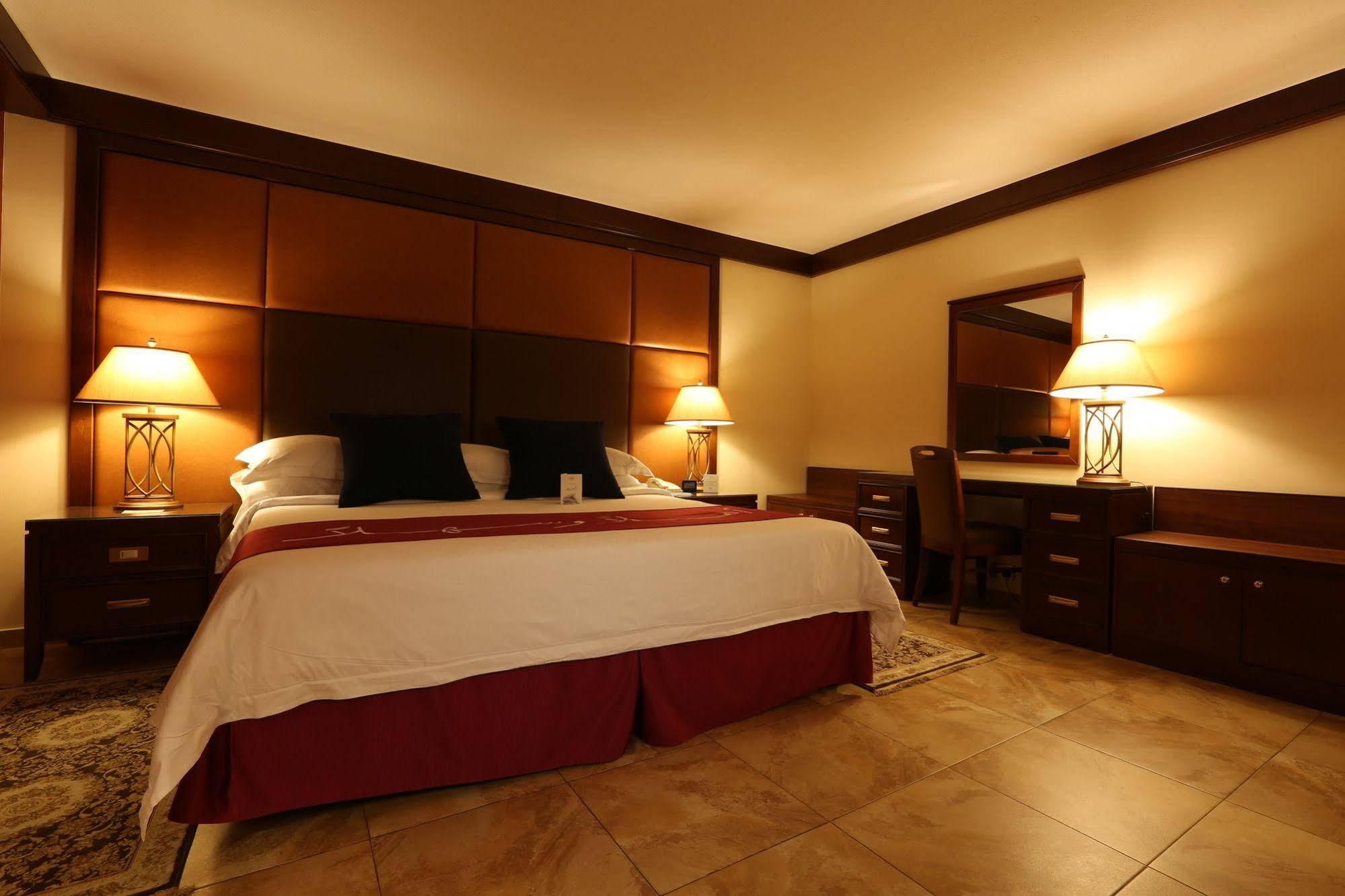 Makarem Ajyad Makkah Hotel Мекка Экстерьер фото The photo shows a well-appointed hotel room with a cozy and inviting atmosphere. It features a large bed with a white comforter and dark pillows, positioned against a wooden headboard. There are two bedside lamps providing soft lighting on either sid