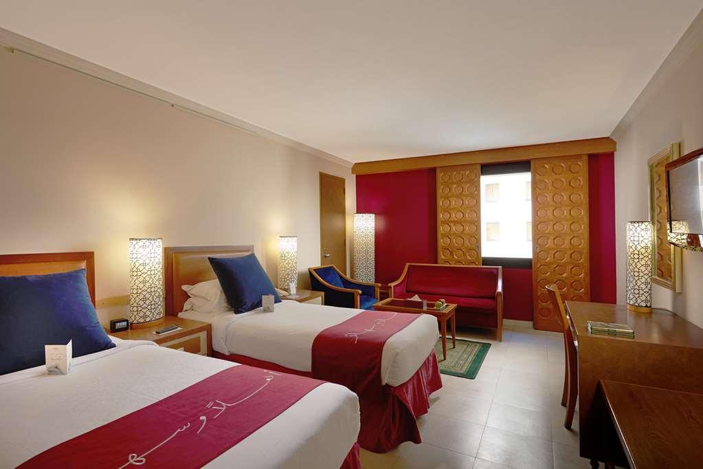Makarem Ajyad Makkah Hotel Мекка Номер фото The photo shows a hotel room featuring two twin beds with red and white bedding. There are decorative lamps on either side of the beds. A small seating area is visible, including a blue chair and a red sofa. There is a wooden desk against one wall, a