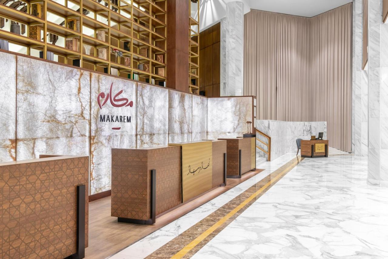 Makarem Ajyad Makkah Hotel Мекка Экстерьер фото The photo shows a modern hotel reception area with a sleek design. The reception desk is made of a combination of wood and stone, featuring a warm color palette. The backdrop is adorned with a marble finish, giving a luxurious feel. Above the desk, t