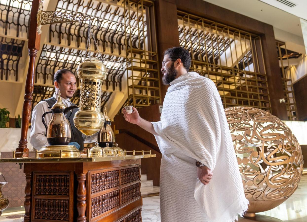Makarem Ajyad Makkah Hotel Мекка Экстерьер фото The photo depicts a scene in a luxurious setting, likely a hotel or a fine dining establishment. In the foreground, a man wearing traditional attire, possibly a white garment, is engaging with a staff member. The staff member is behind a wooden count
