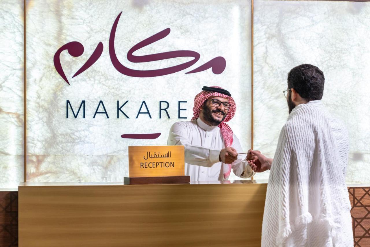 Makarem Ajyad Makkah Hotel Мекка Экстерьер фото The photo shows a reception area with a sign that reads "MAKARE" in both Arabic and English. In the foreground, a staff member wearing traditional attire is smiling and shaking hands with a visitor, who is clad in a white garment, typical for formal 