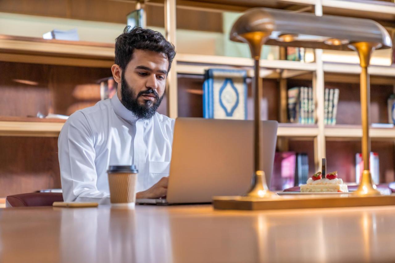 Makarem Ajyad Makkah Hotel Мекка Экстерьер фото The photo shows a man sitting at a desk in a library or a study area. He is wearing a white traditional garment and is focused on working on a laptop. In front of him, there is a cup of coffee and a small cake decorated with strawberries. The backgro