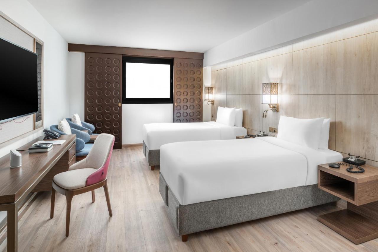 Makarem Ajyad Makkah Hotel Мекка Экстерьер фото The photo shows a modern hotel room featuring two beds with white linens. There is a wooden accent wall with a window above it. Opposite the beds, a wall-mounted television is visible. A stylish wooden desk with a chair is situated on one side of the