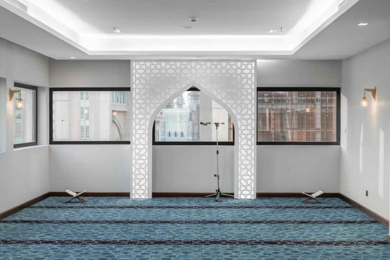 Makarem Ajyad Makkah Hotel Мекка Экстерьер фото The image shows a prayer room or small mosque interior. It features a white decorative wall with an archway that is typical in Islamic architecture. The floor is covered with a blue carpet, arranged with patterned lines, likely indicating prayer spot