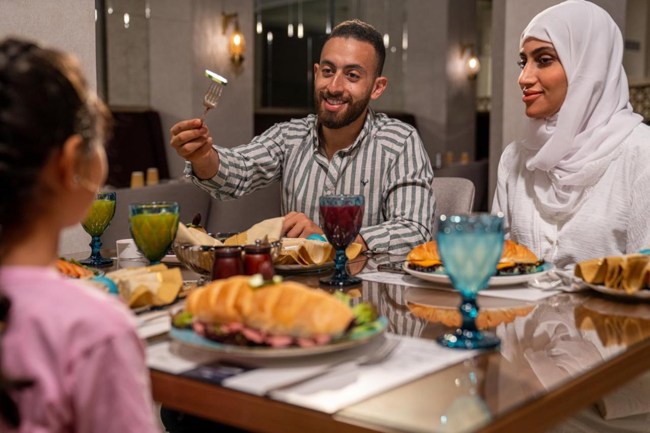 Makarem Ajyad Makkah Hotel Мекка Экстерьер фото The photo shows a dining scene with three people enjoying a meal together. A man is seated at the table, smiling and holding a utensil, possibly ready to serve or share food. He is dressed in a striped shirt. Across from him is a woman wearing a whit