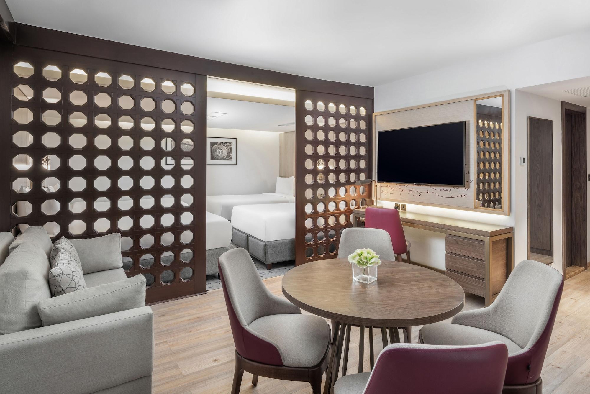 Makarem Ajyad Makkah Hotel Мекка Экстерьер фото The photo shows a modern hotel room or suite. It features a living area with a comfortable gray sofa and a round wooden dining table surrounded by upholstered chairs. A large flat-screen TV is mounted on the wall opposite the seating area. There's a 
