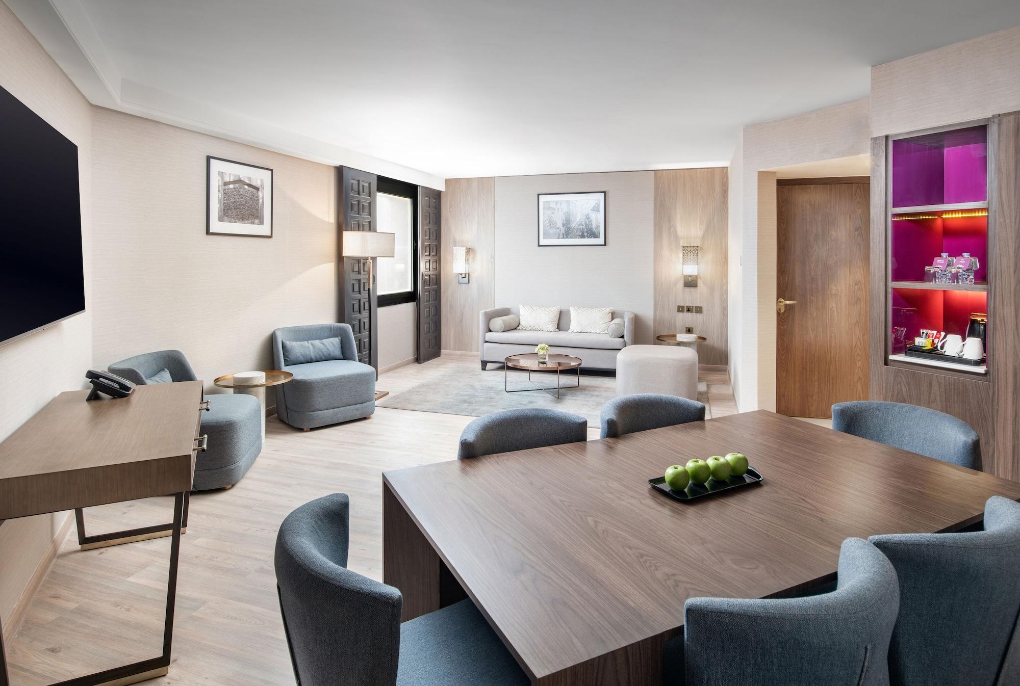 Makarem Ajyad Makkah Hotel Мекка Экстерьер фото The photo shows a modern and elegantly furnished living area. It features a spacious layout with a comfortable seating arrangement that includes a sofa and armchairs. There is a large wooden dining table with several chairs around it. The walls are a
