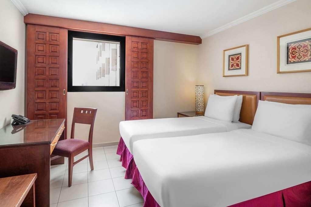 Makarem Ajyad Makkah Hotel Мекка Номер фото The photo shows a hotel room with two beds, each covered with white bedding. There is a wooden desk and a chair near the beds. The room features a window with a wooden frame and a decorative lamp beside the beds. On the wall, there are two framed art