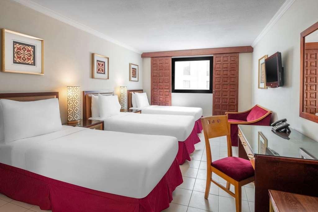 Makarem Ajyad Makkah Hotel Мекка Номер фото The photo shows a hotel room featuring two twin beds with white linens and red bed skirts. There are decorative elements on the walls, including framed artwork. A large window provides natural light, while a desk and a chair are positioned next to it