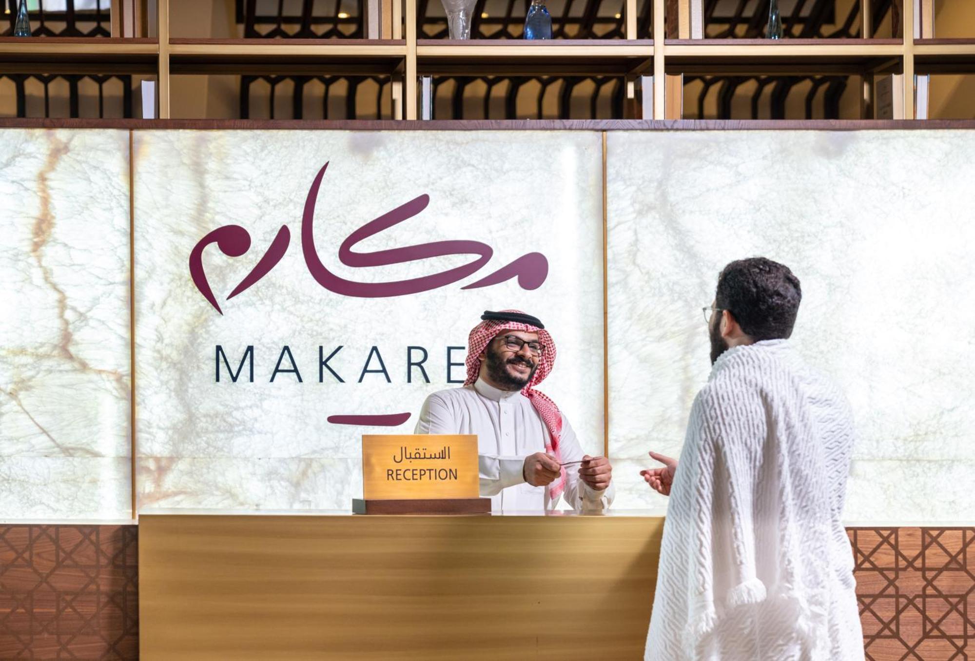 Makarem Ajyad Makkah Hotel Мекка Экстерьер фото The photo depicts a reception area in a hotel or establishment named "Makarem." In the image, a receptionist, dressed in traditional attire, is smiling and interacting with a guest who is wearing a white robe, commonly known as a thobe or ihram. The 