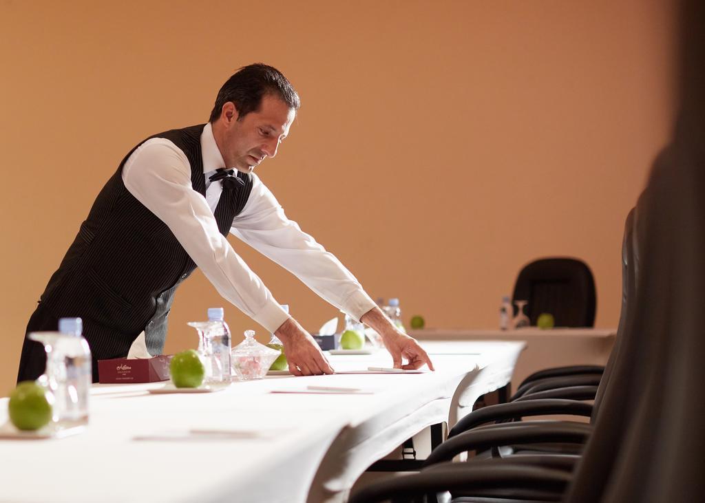 Makarem Ajyad Makkah Hotel Мекка Экстерьер фото The image shows a waiter or service staff member in a formal attire, which includes a vest and bow tie. He is preparing a long table, likely for an event or meeting. The table is set with items such as water bottles and green apples, and there are se