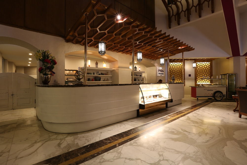 Makarem Ajyad Makkah Hotel Мекка Экстерьер фото The photo shows a modern reception area or lobby of a hotel or restaurant. The focal point is a curved front desk made of white material, featuring a glass display case that showcases various items. Above the desk, there is a decorative wooden struct